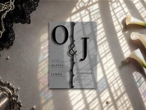 The wedding invitation lies on a neutral background next to white calla lilies, a beaded lace trim, and window shadow patterns.