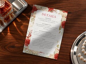 Details card for a wedding with red floral borders, resting on a wooden surface.