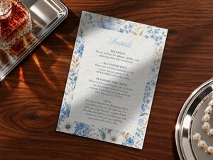 A wedding details card decorated with blue wildflowers rests on a wooden table next to a crystal decanter and a silver tray.