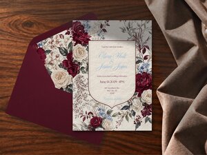 A wedding invitation card with burgundy and white floral borders sits partially inside a matching dark red envelope on a wooden surface.