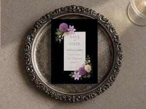 A black wedding save-the-date card with purple floral corners rests on an ornate silver tray.