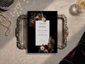 A Save the Date wedding card with a black background and white text, decorated with brown and burgundy floral arrangements, resting on a silver tray.