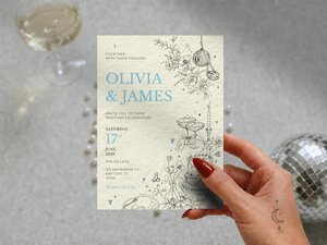 A hand holds up the Olivia and James wedding invitation card against a textured background with a glass of champagne and disco ball.