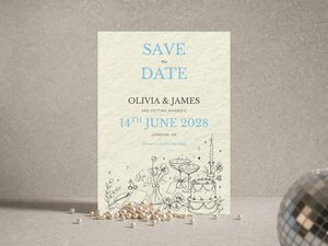 A Save the Date card featuring blue text and line art sits on a surface with scattered pearls and a disco ball.