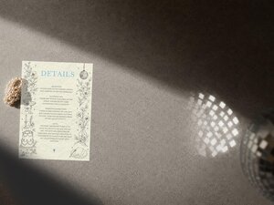 A wedding details card is placed on a grey textured surface next to a pearl bracelet and light reflecting from a disco ball.