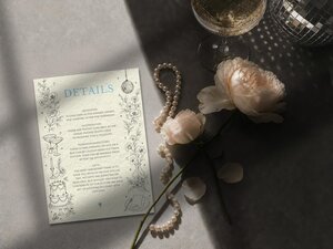 A details card with blue and black ink sits beside pale pink roses, a string of pearls, and a glass of champagne.