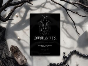 A black wedding invitation featuring two facing skeletons, surrounded by dark branches, pearls, and a crystal.