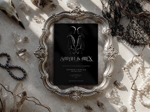 The invitation lies inside an ornate silver vintage frame on a surface decorated with pearls, crystals, and dark lace.