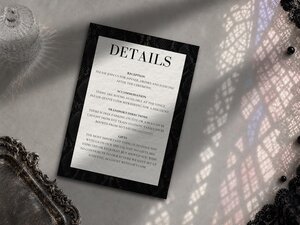 A wedding details card with a dark patterned border rests on a surface with shadow patterns, next to a silver tray.