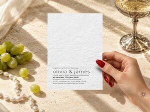 A hand holding a textured white wedding invitation card surrounded by green grapes, pearls, and a coupe glass on a neutral surface.