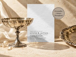 A textured white wedding invitation card displayed next to a silver pedestal bowl and a silver tray on a beige stone background.