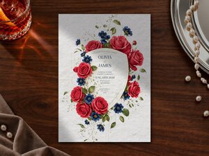 A wedding invitation featuring red roses and blue flowers on textured white paper, displayed on a wooden table with a glass of amber liquid and pearls.