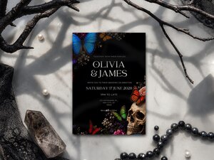 A wedding invitation with a skull and butterfly design lying flat on a marble surface decorated with black beads, crystals, and dark branches.