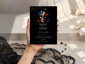 A person holding a Save the Date card with a skull and butterfly design over a decorative tray and pearl strands.