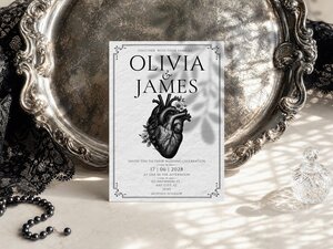 A wedding invitation resting in front of an ornate silver tray, with black lace and pearl beads on the surface.