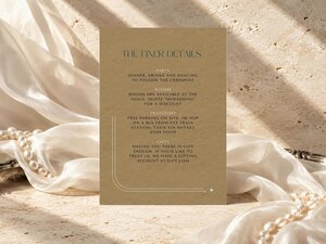 Gold finer details card with dark teal text resting on a stone surface among white draped fabric and strands of pearls.