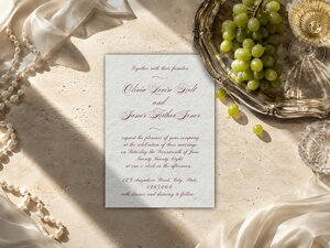 A wedding invitation template sits flat on a textured beige surface near a silver tray of green grapes and strands of pearls.