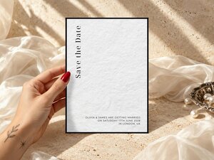A hand with red nail polish holds a save-the-date card for Olivia and James against a textured stone backdrop with white fabric.