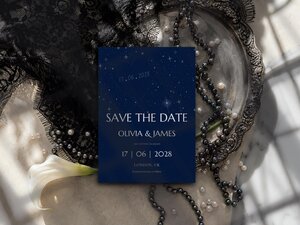 A dark blue celestial wedding invitation lies on a silver tray surrounded by black lace and pearl strands.