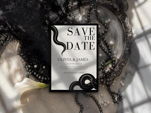 A save the date card with black snake illustrations is displayed on a surface alongside a silver tray, pearl beads, black lace, and a white calla lily flower.