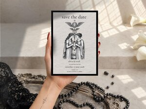 A hand holds a Save the Date card featuring a tarot-style illustration of two women, displayed against a light surface with black lace, pearls, and white calla lilies.