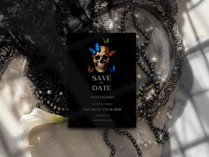 A black save the date card featuring a human skull with colorful butterflies, resting on a stone surface with black lace, pearls, and a white flower.