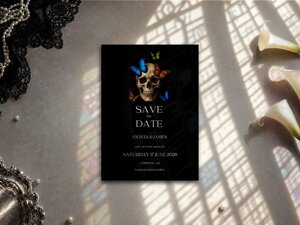 The save the date card lies on a textured stone surface illuminated by patterned window light, surrounded by white flowers and a silver tray.