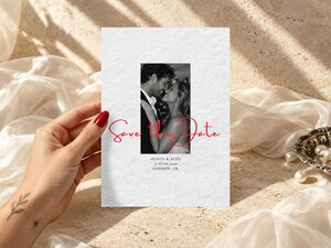 A hand holds a save the date card featuring a black and white couple photo and red script lettering.