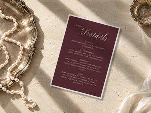 A Details card for a wedding is placed on a stone surface beside a silver tray with pearls and a small glass dish.