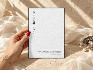 A hand with red nail polish holding a save the date card against a beige textured background with white draped silk fabric and pearls.