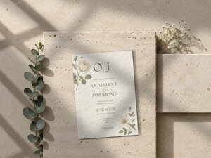 A botanical wedding invitation lies on a stacked stone surface alongside a sprig of eucalyptus and small white flowers.