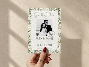 A hand holds a save the date card featuring a black and white couple photo, floral border, and Lake Como Italy stamp.