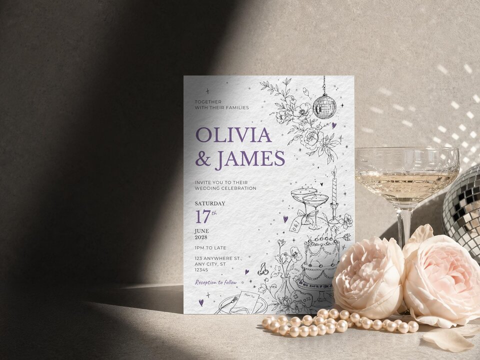 A wedding invitation with purple text and line art illustrations of a cake, disco ball, and game controller stands upright next to a champagne glass and pink roses.