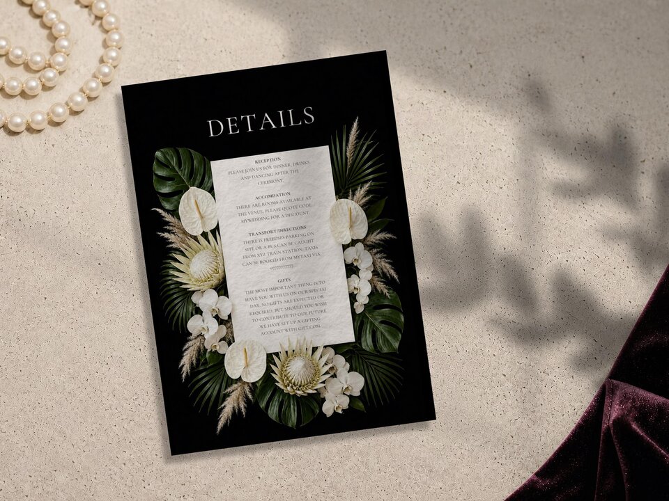 A wedding details card template with a tropical floral border on a black background, resting on a stone surface beside dark fabric.