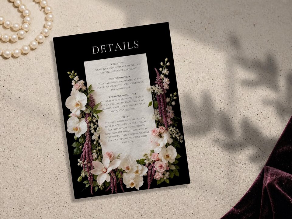 A wedding details card on a black background with white floral arrangements on both sides, lying on a beige stone surface beside a pearl necklace and dark velvet fabric.