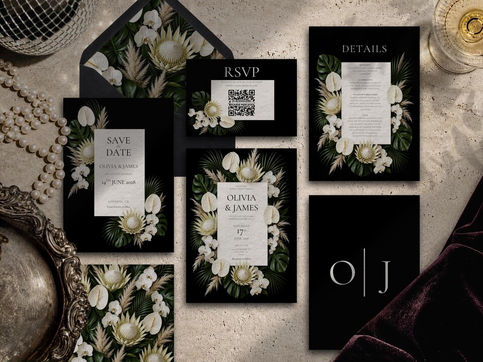 A flat lay of a complete black and white tropical floral wedding invitation suite displayed on a textured stone surface with pearls and an open envelope.
