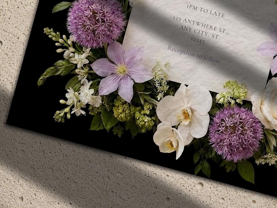 Close-up angled view of a wedding invitation card on a black background, decorated with purple alliums, white orchids, and lilac clematis flowers.
