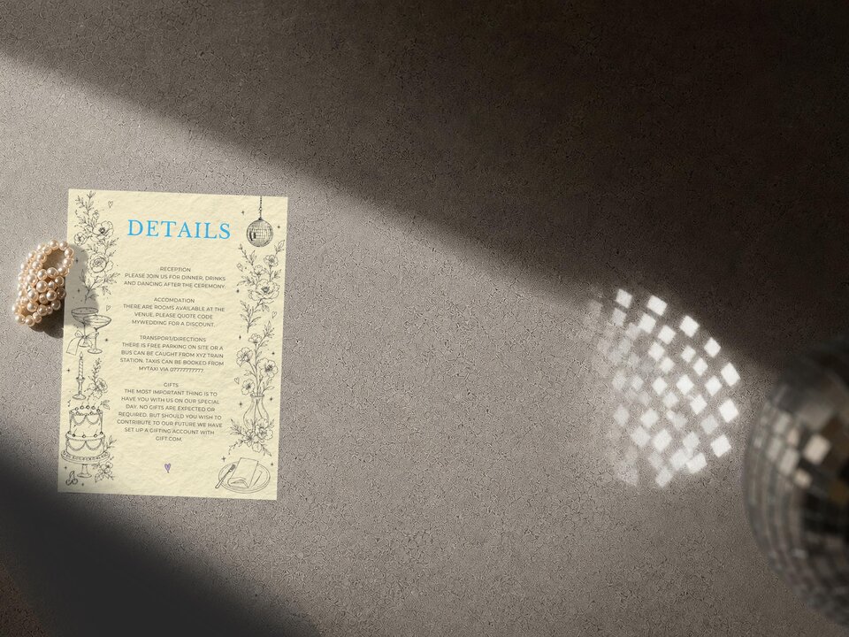 A wedding details card with blue typography and floral line art resting on a textured gray surface beside pearls.