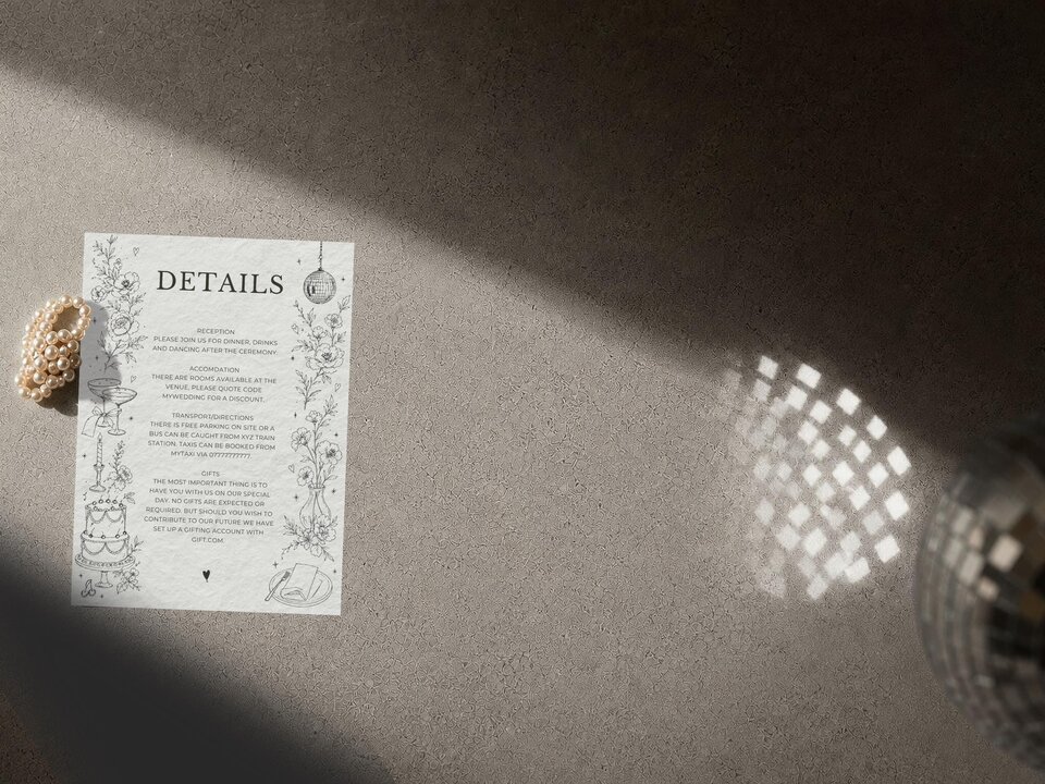 A wedding details card resting on a gray textured surface with a pearl bracelet on the left and a glittering disco ball on the right.