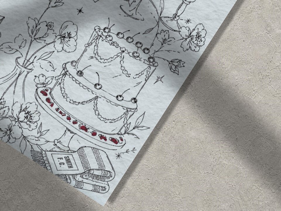 Close-up of the illustrated wedding stationery featuring a multi-tiered cake, flowers, and a football scarf on a textured surface.