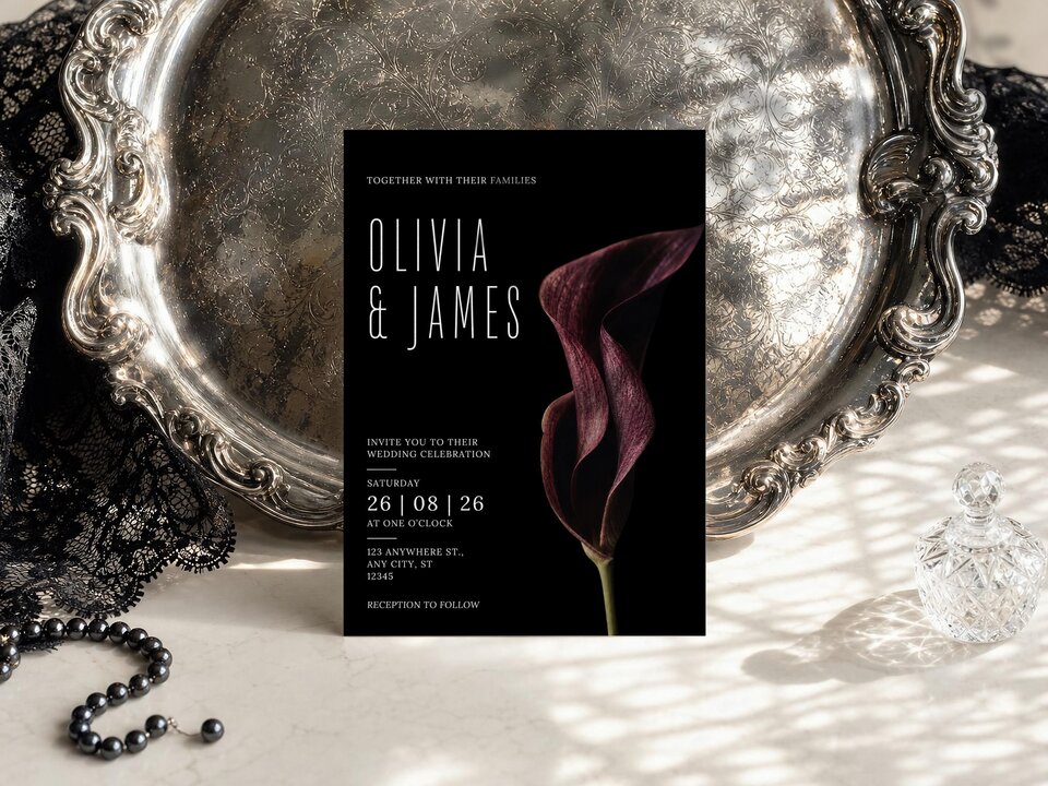 A wedding invitation with a dark burgundy calla lily graphic is displayed on a decorative silver tray next to a black lace cloth.