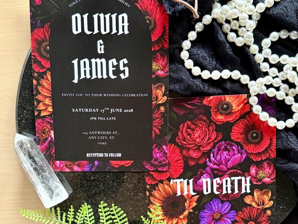 A flat lay of two wedding stationery cards featuring dark floral prints, accompanied by white pearls, a crystal point, and a green fern frond.