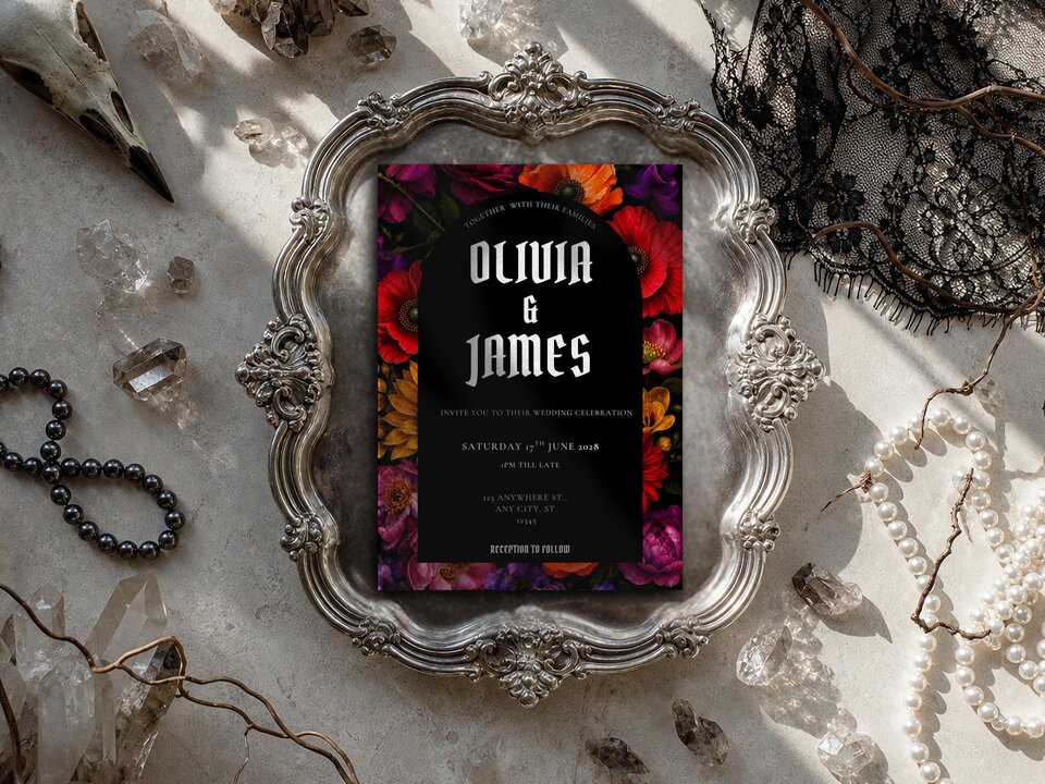 A gothic wedding invitation is displayed inside an ornate silver frame on a surface decorated with crystals and lace.