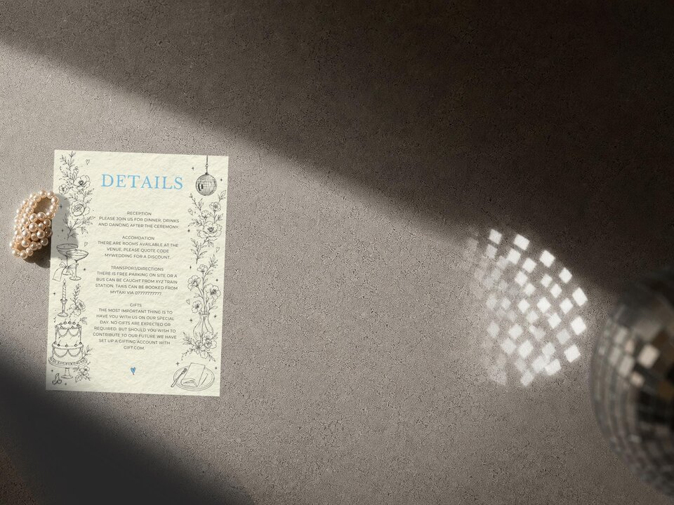 A wedding details card is placed on a grey textured surface next to a pearl bracelet and light reflecting from a disco ball.