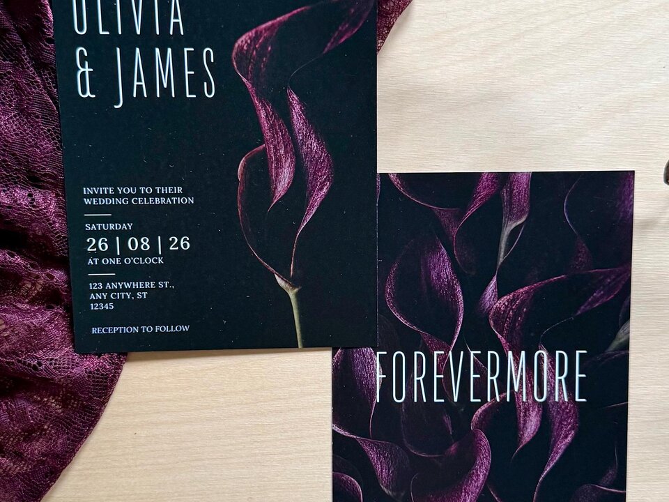Two wedding stationery pieces with a dark burgundy calla lily design rest on a wooden surface beside burgundy fabric and a dark branch.