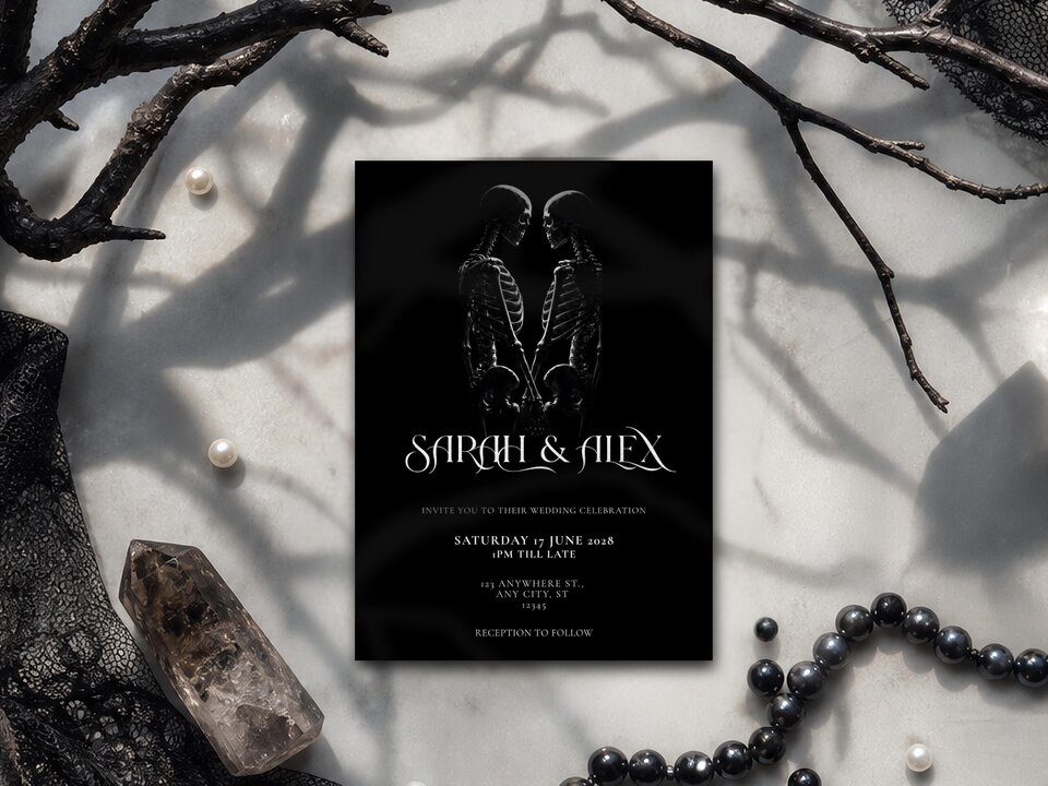 A black skeleton wedding invitation rests on a white marble surface surrounded by pearls, a crystal point, dark lace, and bare branches.