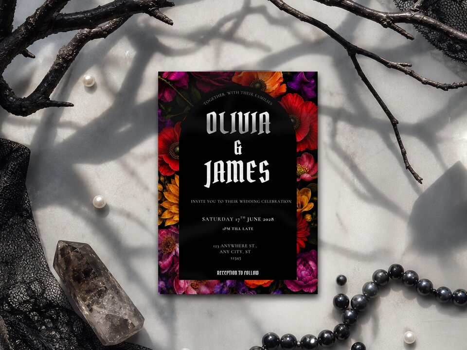 A gothic floral wedding invitation rests on a marble surface among dark branches, crystals, and black beads.