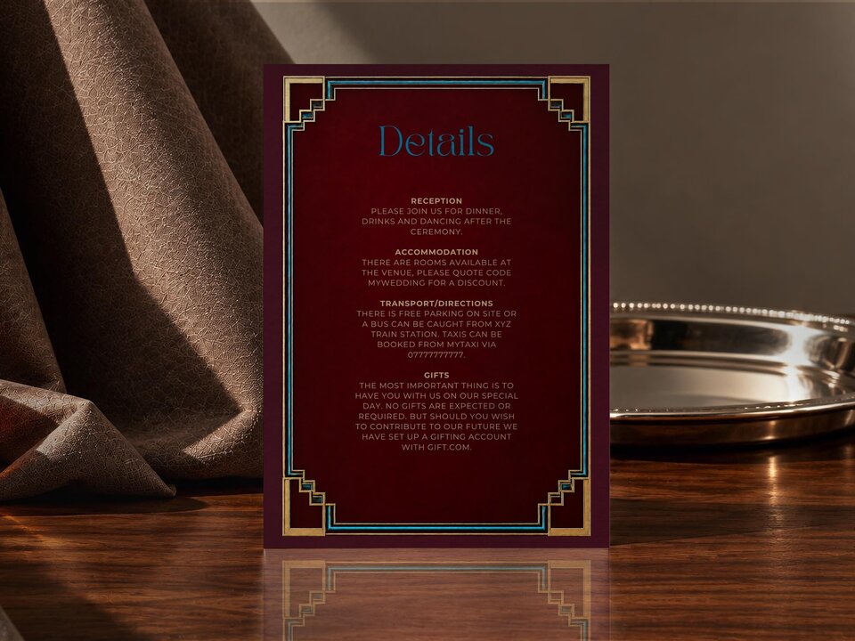 A wedding details card with art deco borders is placed next to draped fabric and a silver tray.